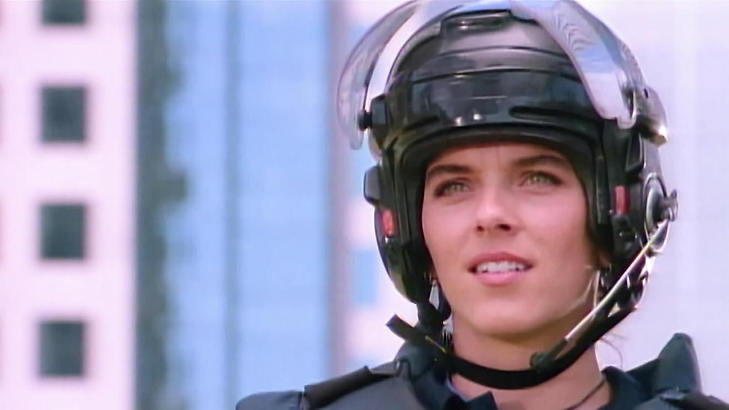 Eine Frau mit schwarzem Helm und Uniform. Sie schaut nach oben und zur Seite. Gebäude sind im Hintergrund.