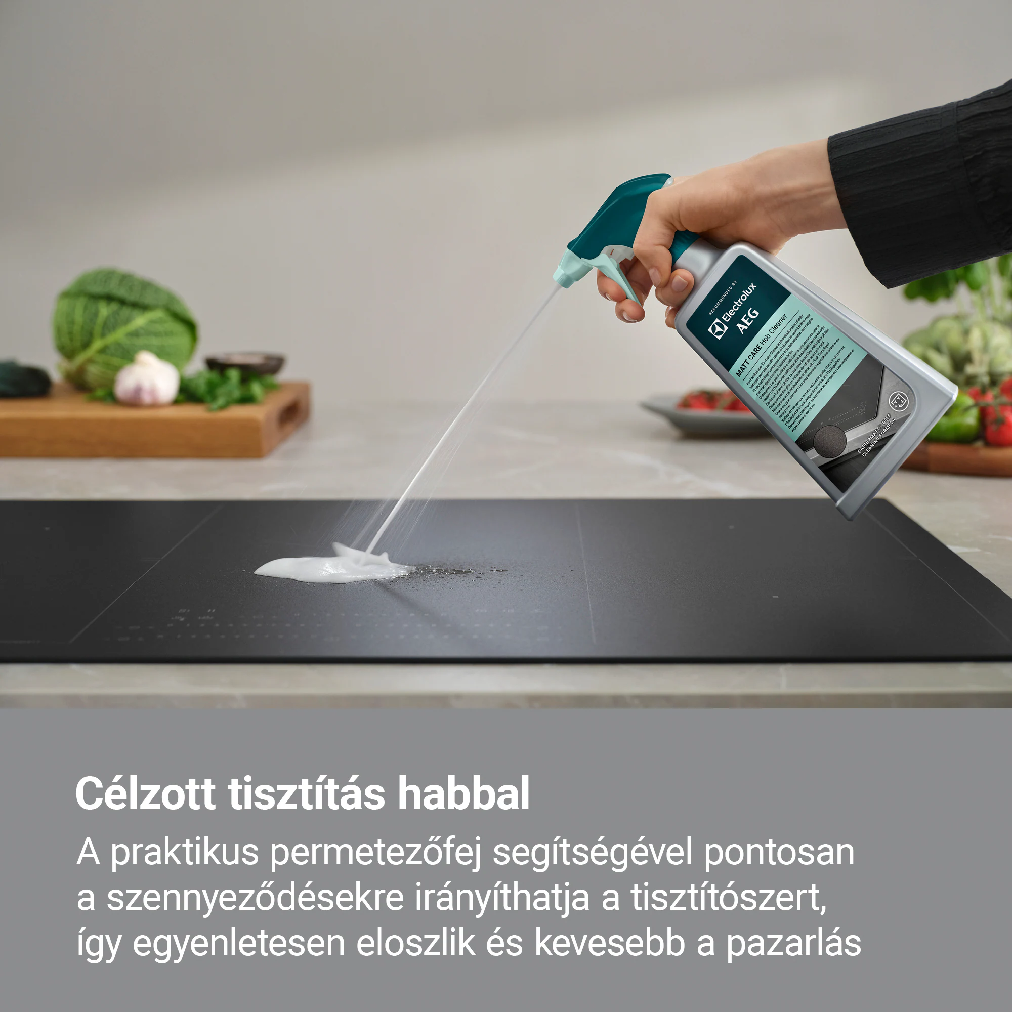 Egy kéz tisztítóhabot permetez egy fekete tűzhelyre. Zöldségek a háttérben. Egy AEG tisztítószeres flakon látható.