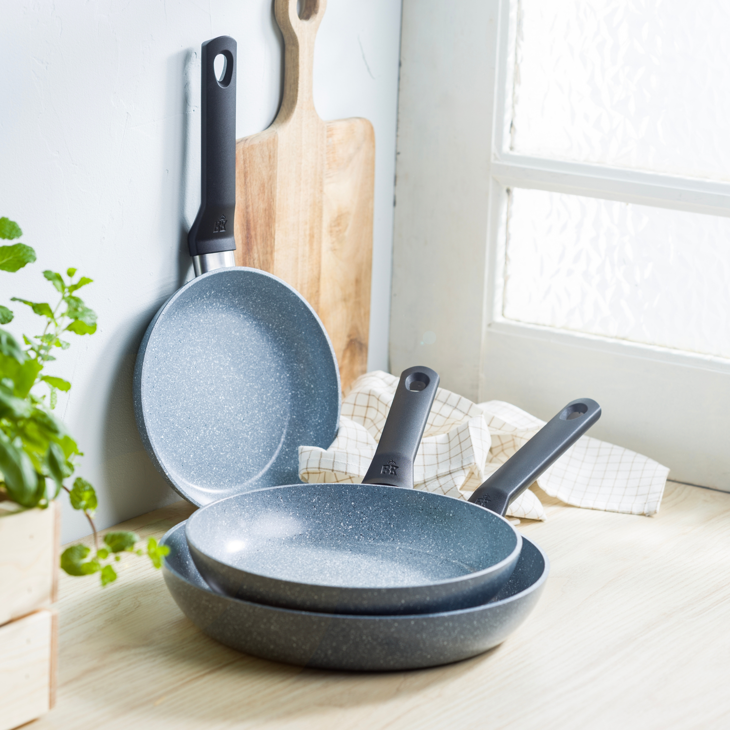 Three grey pans with black handles are on a wooden surface. A wooden cutting board and a window are in the background.