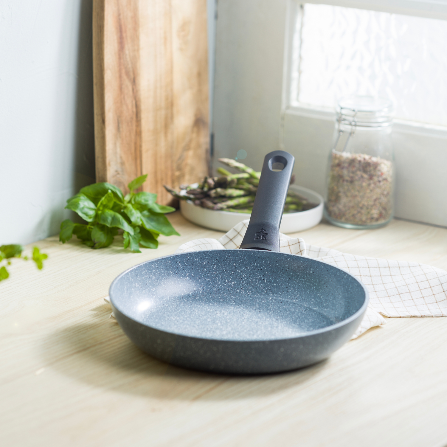 A gray speckled frying pan on a wooden counter. A wooden cutting board, basil, asparagus, and a jar are in the background.