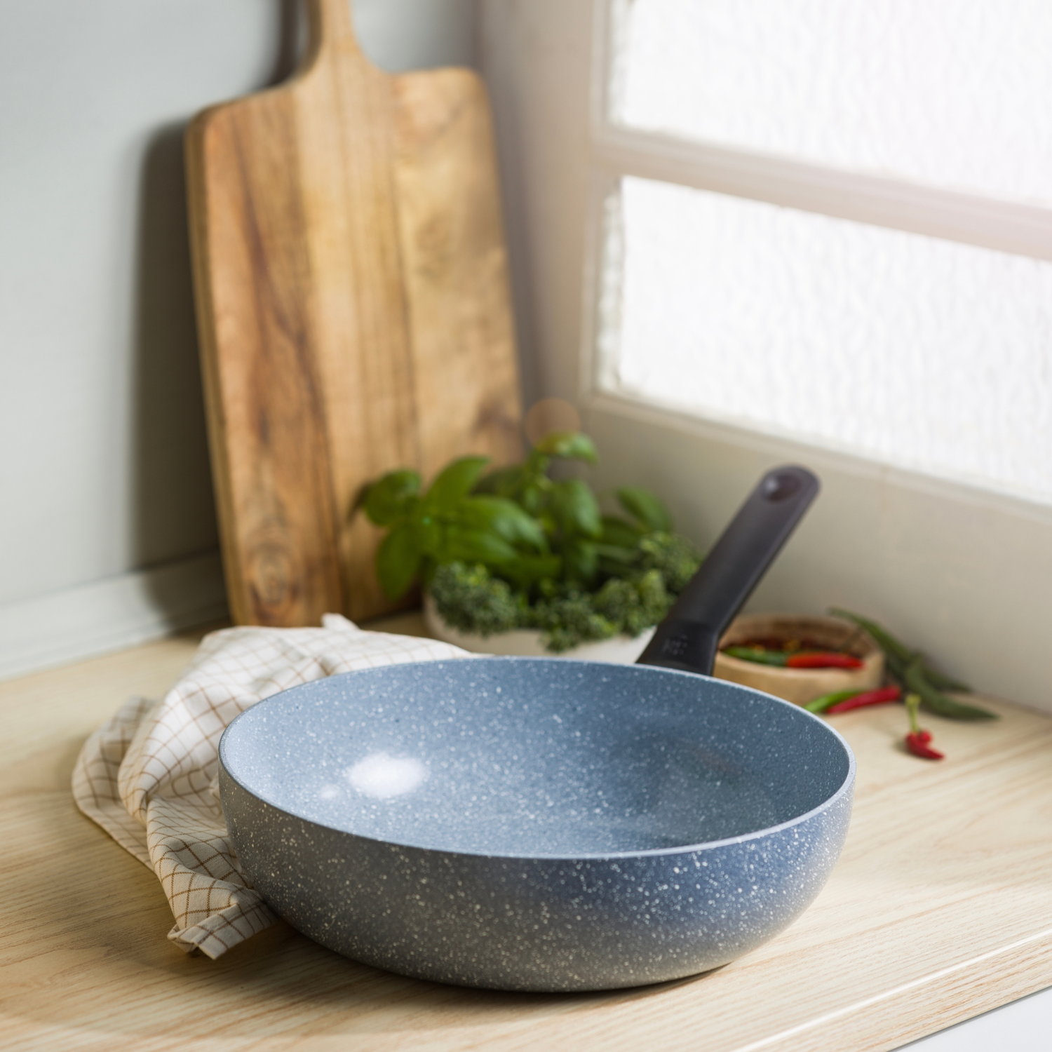 A gray speckled pan sits on a wooden counter. A cutting board, basil, and peppers are in the background.