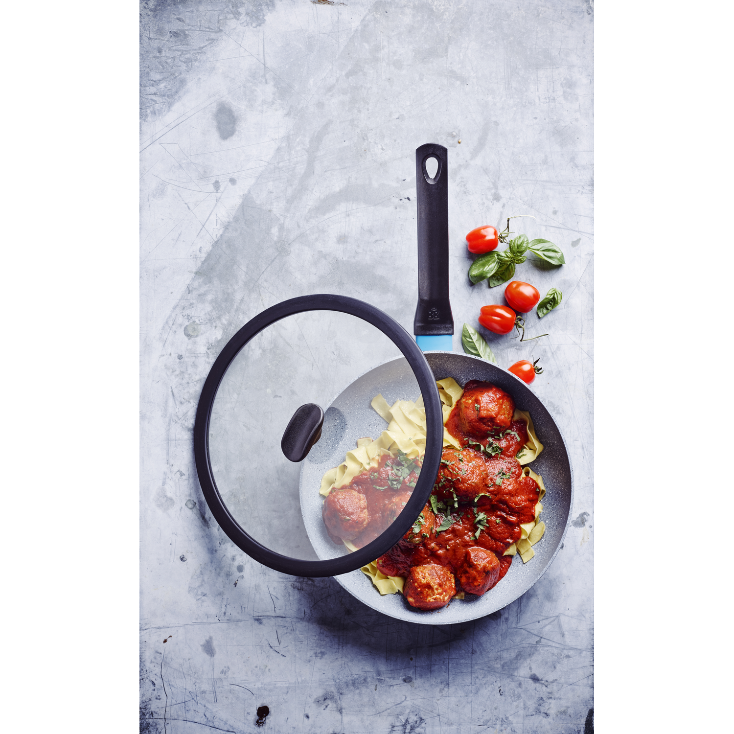 A pan with meatballs and pasta, a glass lid, tomatoes, and basil on a gray surface. The pan has a black handle.