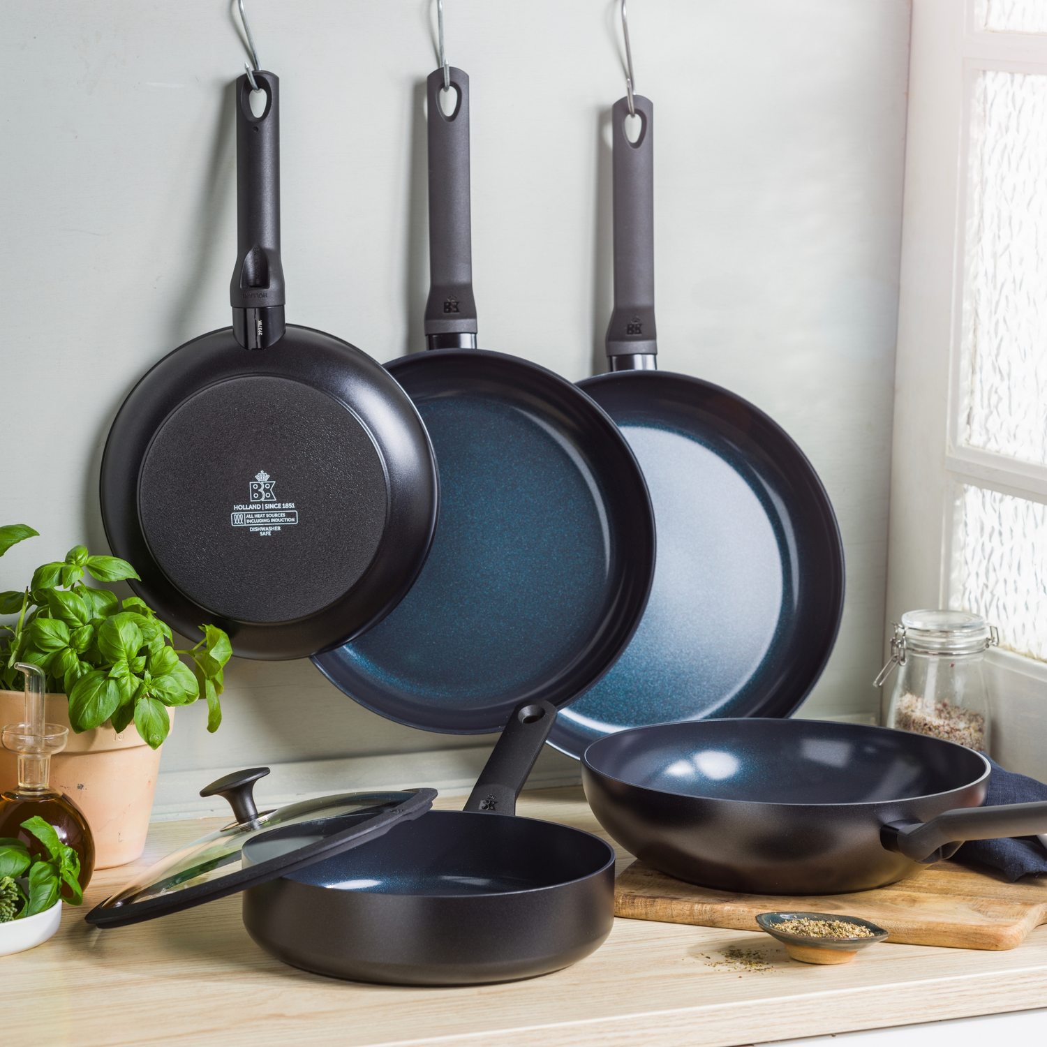 Several black frying pans and a wok. Pans hang on hooks. Basil, olive oil, spices, and a wooden cutting board are on a wooden surface.