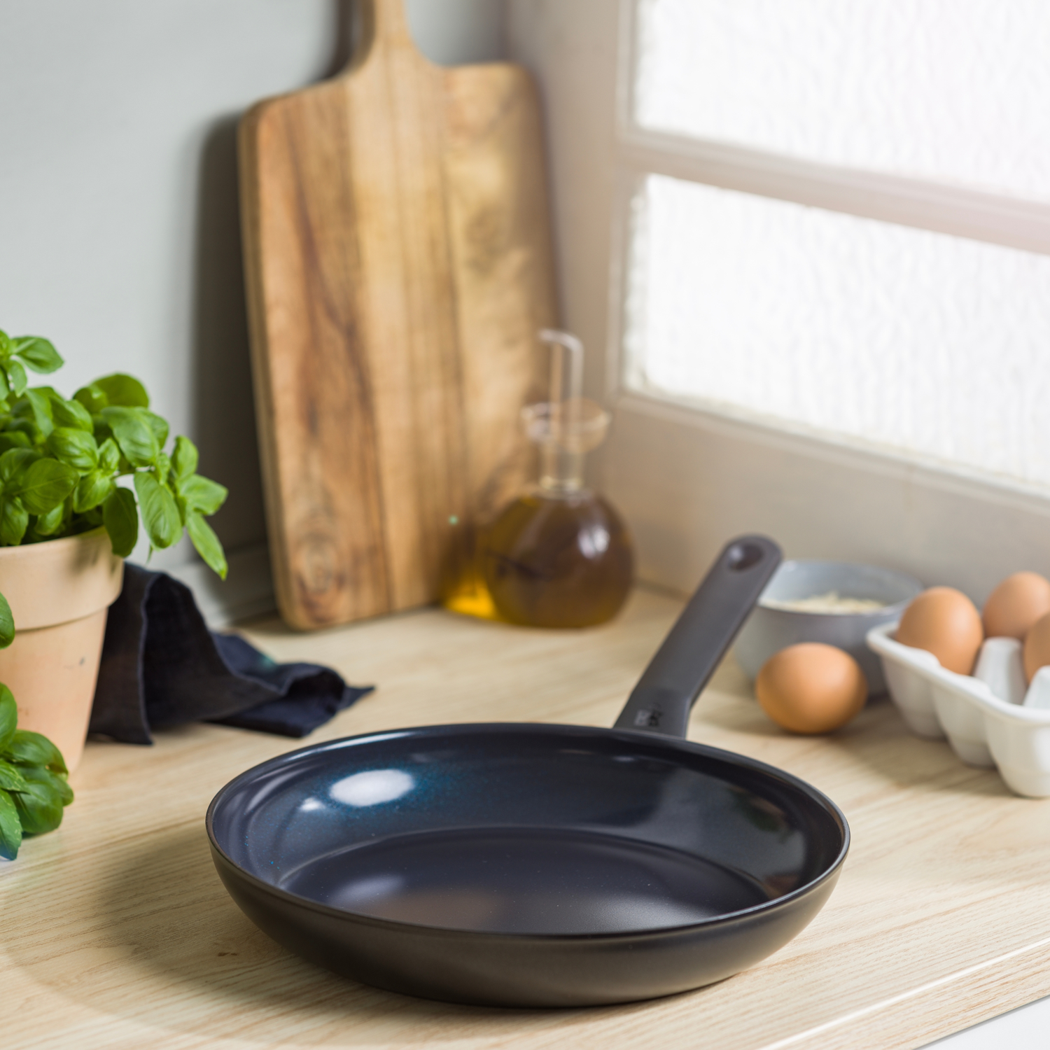 A black frying pan sits on a wooden counter. Eggs, herbs, and oil are visible in the background near a window.