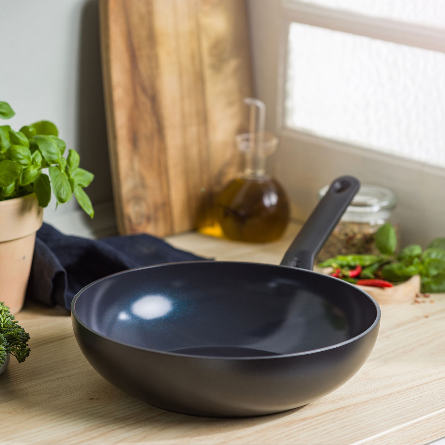 A black pan sits on a wooden surface, with a cutting board, oil bottle, and herbs in the background. A plant and vegetables are on the left.