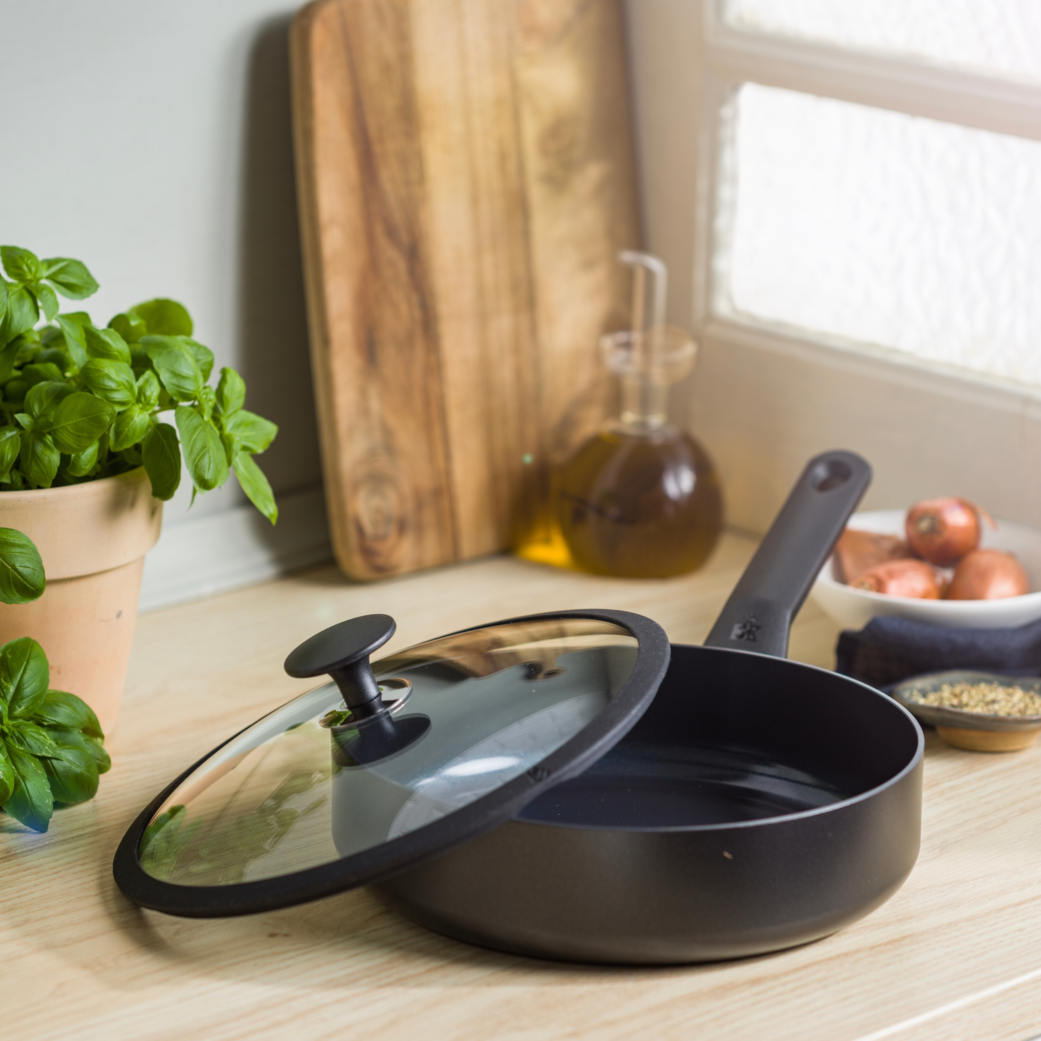A black frying pan with a glass lid sits open on a wooden counter. Basil in a pot, cutting board, and onions are also present.