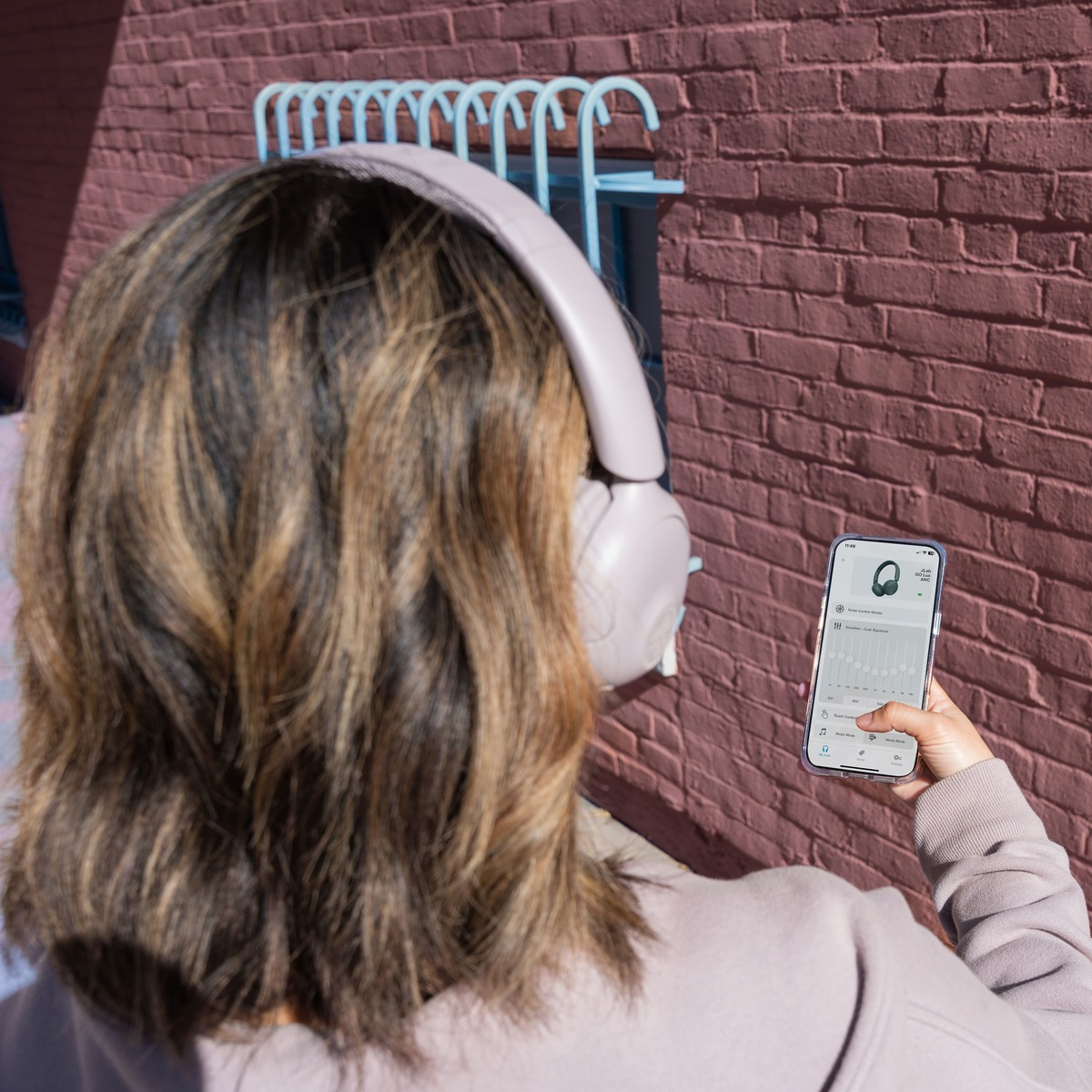 Person mit Kopfhörern, die ein Telefon benutzt. Rote Backsteinmauer im Hintergrund. Kopfhörer sind hellviolett, Telefon in der Hand.