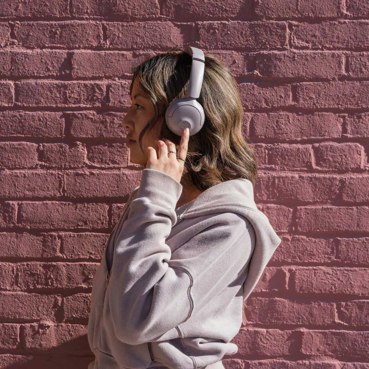 Eine Person mit Kopfhörern und Kapuzenpullover steht vor einer rosa Backsteinmauer und passt die Kopfhörer an.