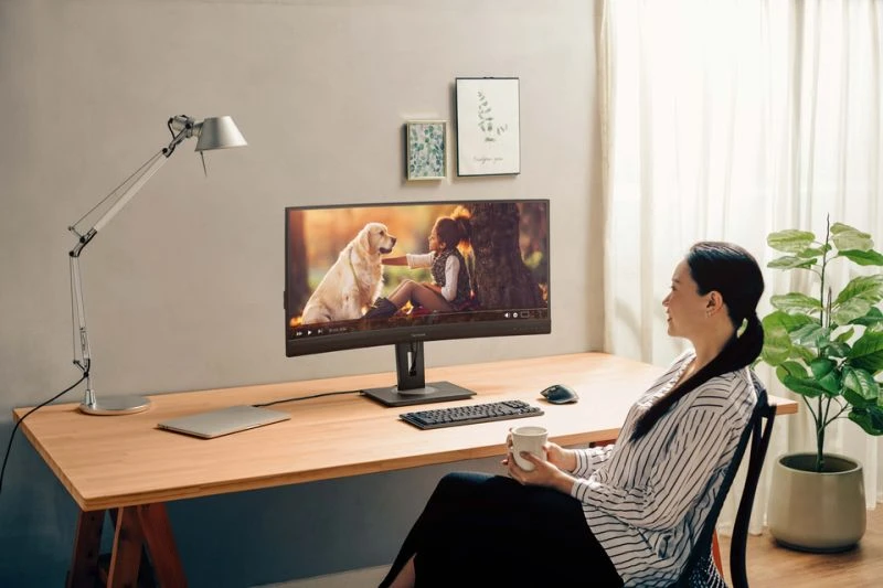 Nő monitort néz. Asztal laptop, billentyűzet és bögre. Növények és fali művészet látható.