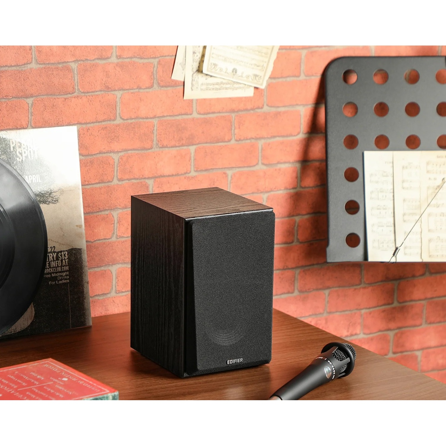 Un altoparlante nero su un tavolo di legno con un microfono e un disco. Un muro di mattoni sullo sfondo.