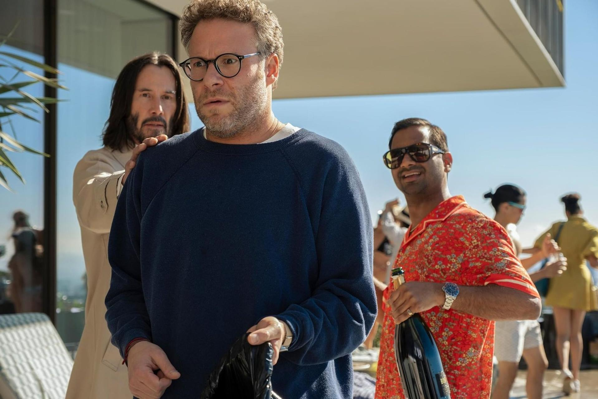 Drei Männer in sonniger Umgebung. Einer trägt Brille und einen blauen Pullover. Andere stehen in der Nähe.
