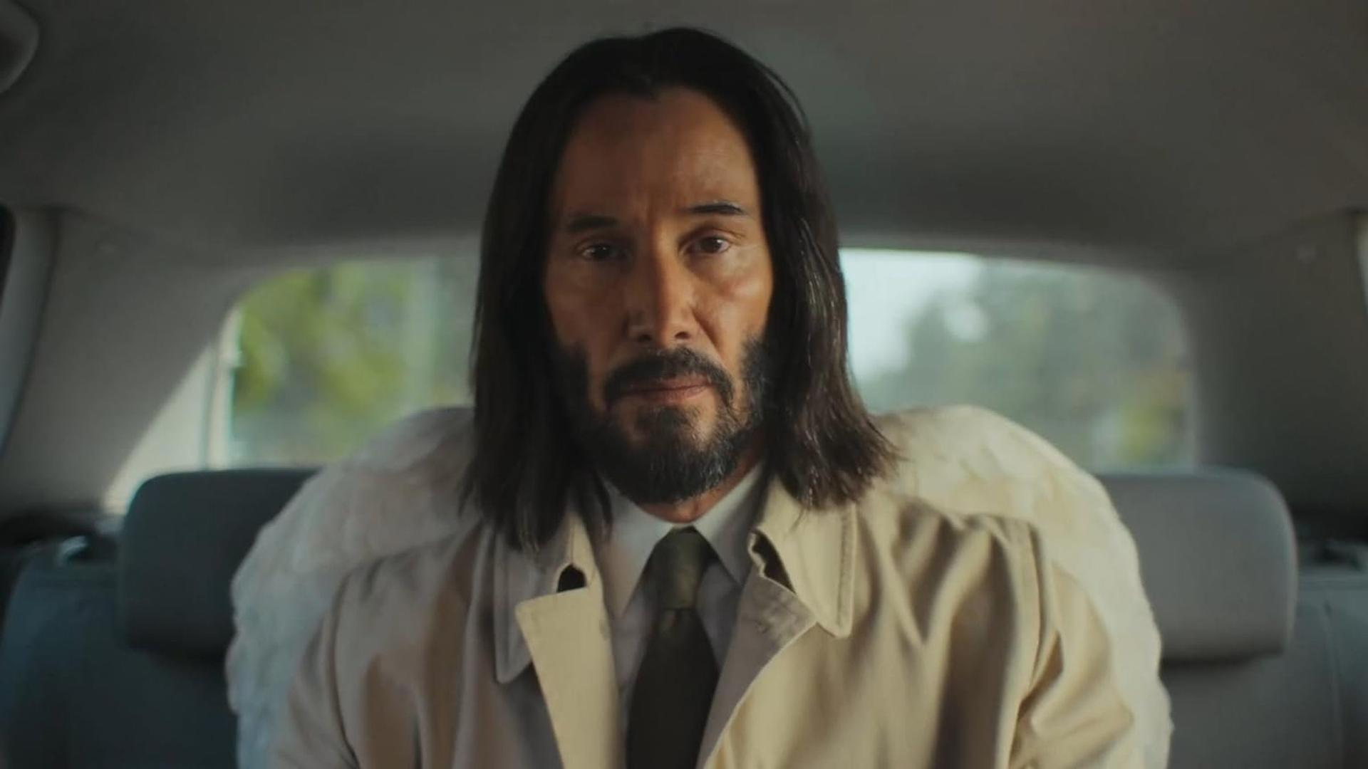 Keanu Reeves im Auto, Blick in die Kamera. Trägt einen beigen Mantel und Krawatte. Hintergrund zeigt das Auto und Bäume.