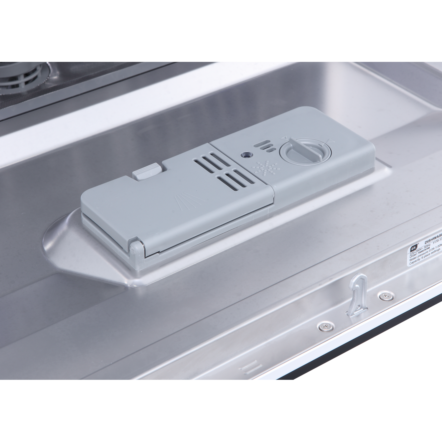 Inside a dishwasher, a grey detergent dispenser is visible. It's rectangular with a hinged lid. The interior is stainless steel.