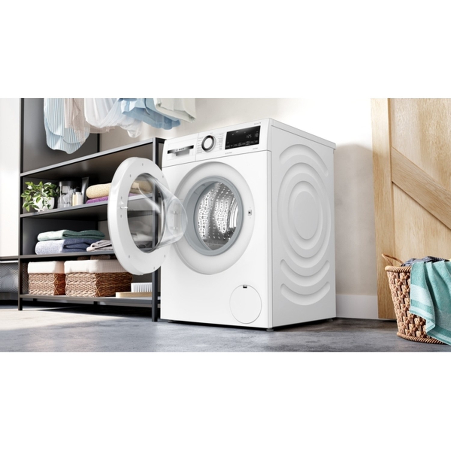 Een witte wasmachine met een open deur. Kleding hangt erboven en is op planken gevouwen. Een mand met handdoeken staat rechts.