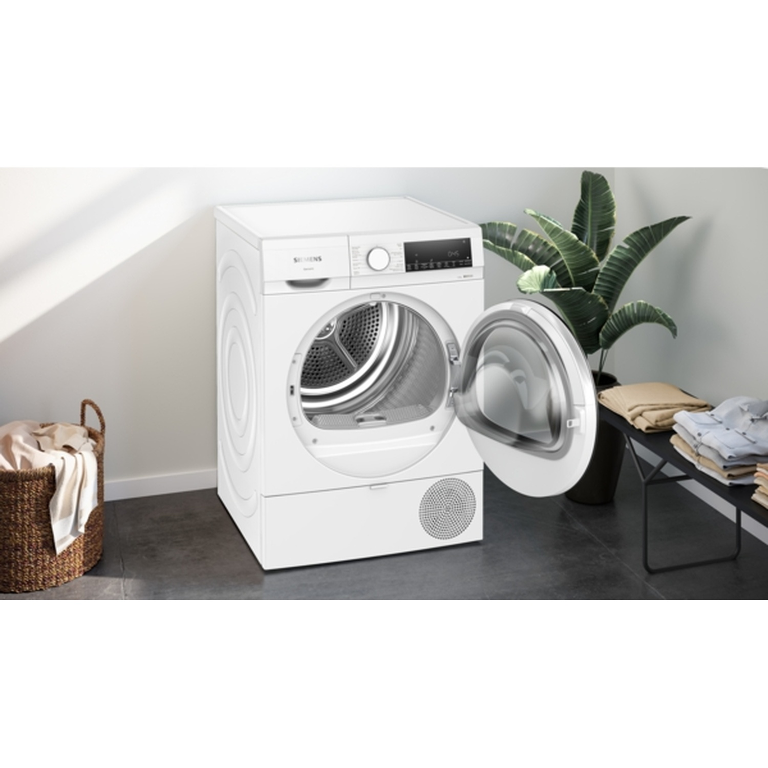 A white dryer with an open door. A wicker basket with clothes is on the left. A plant and clothes are on the right.