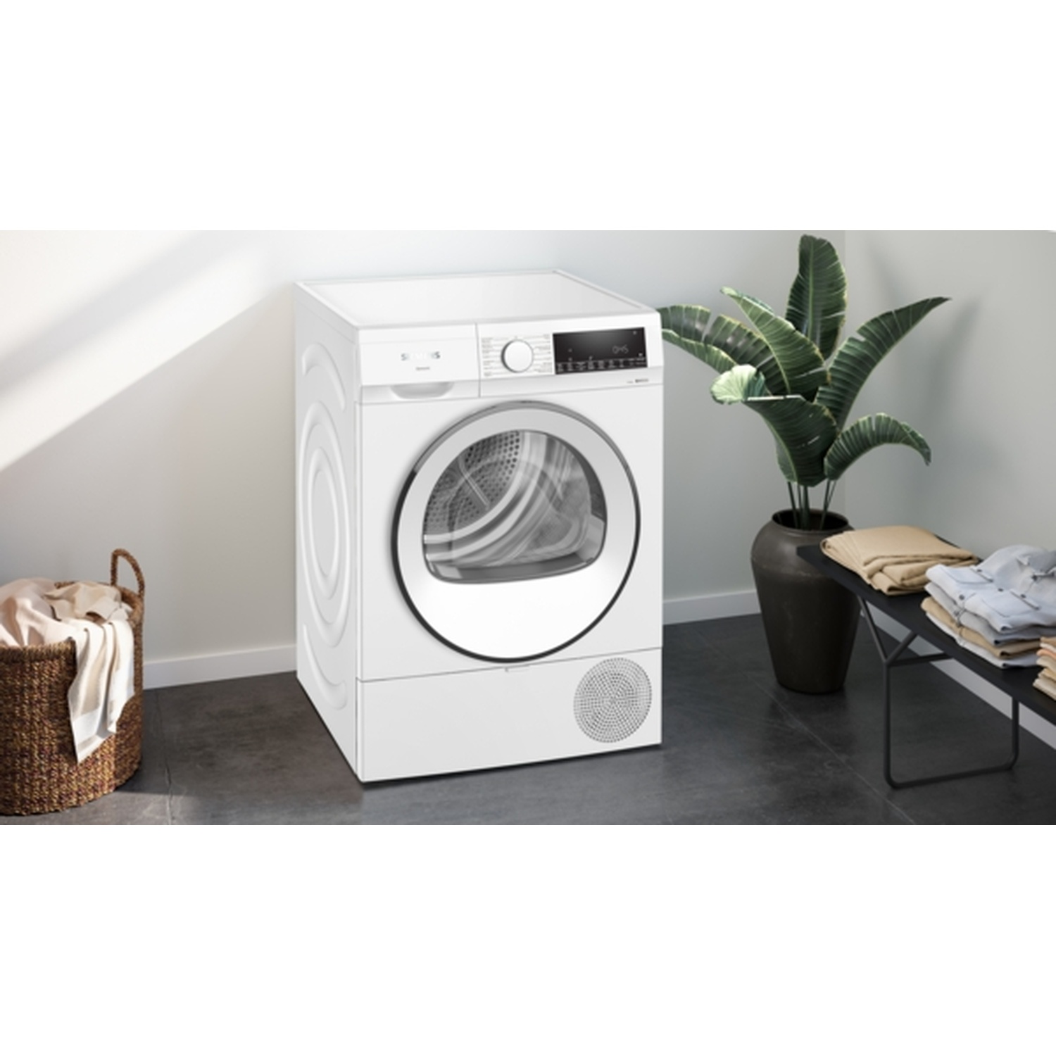A white dryer stands near a laundry basket and a plant. Folded clothes sit on a black bench. The scene is indoors.