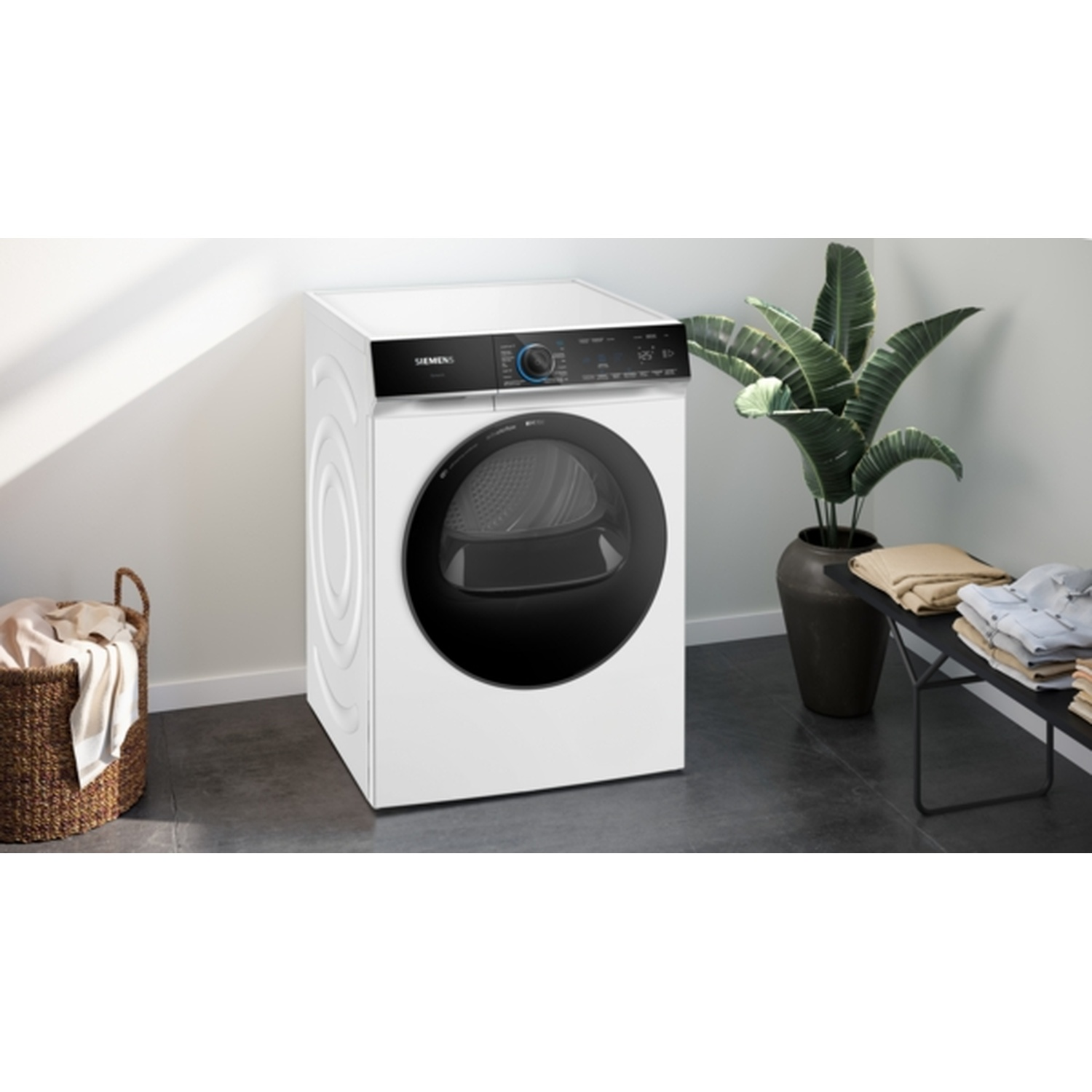 A white dryer with a black door stands in a laundry room. A basket of clothes and potted plant are visible.
