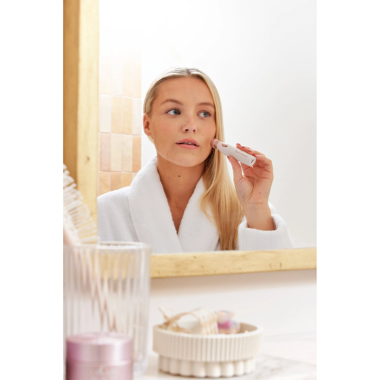 Blonde woman in white robe uses a facial epilator in front of a mirror. Bathroom items are visible in the foreground.