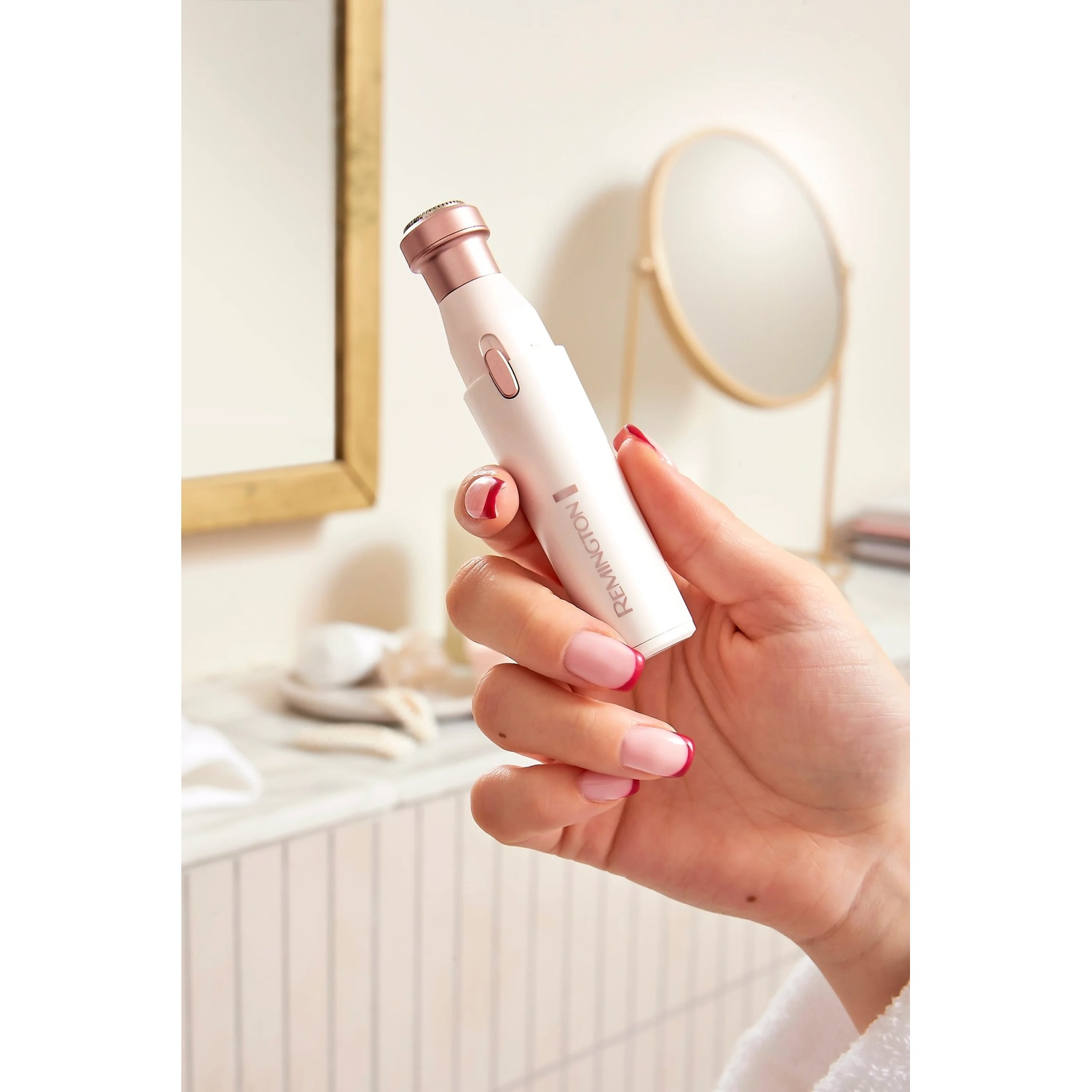 A hand holds a white and rose gold Remington epilator. The hand has manicured nails. A bathroom mirror is in the background.