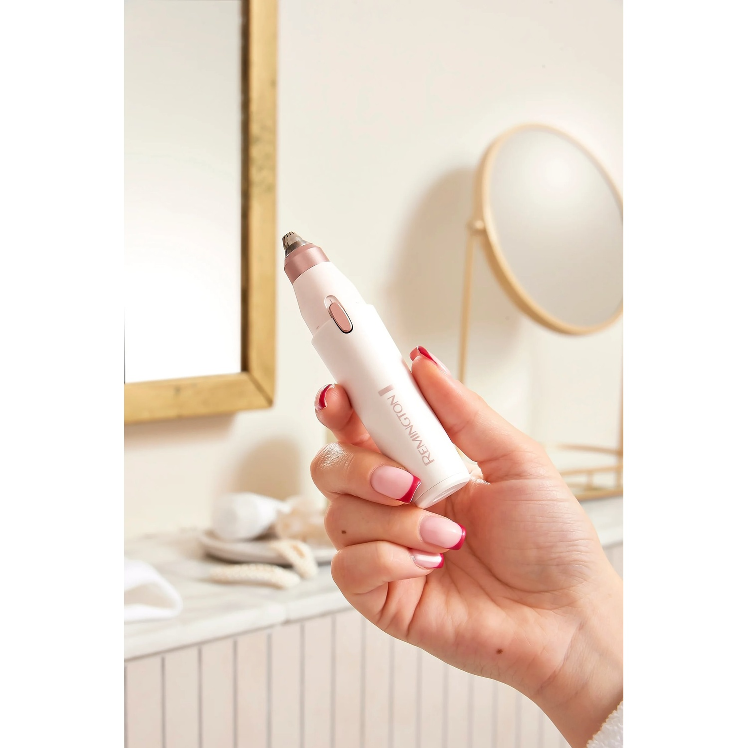 A hand holds a white Remington nose trimmer. The hand has french tip nails. A mirror and bathroom items are in the background.
