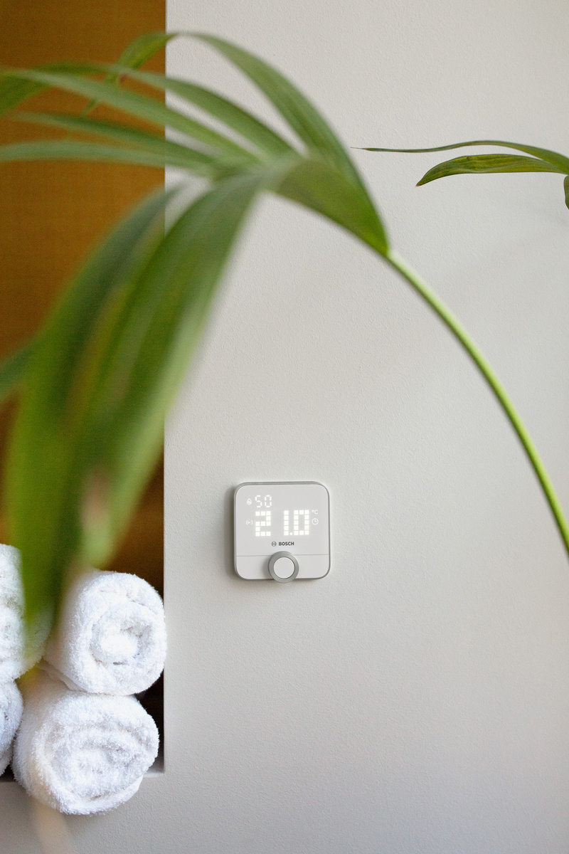 Ein weißes Thermostat zeigt 21 Grad an. Gerollte Handtücher sind links, grüne Blätter im Vordergrund, vor weißer Wand.