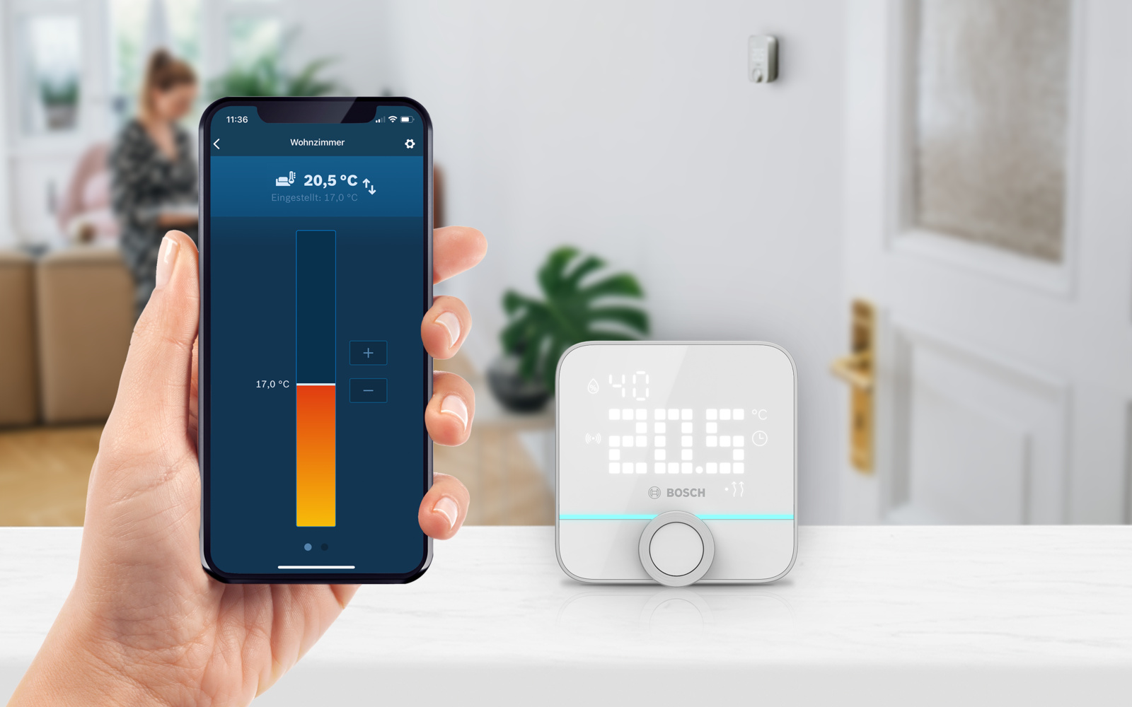Eine Hand hält ein Telefon, das die Raumtemperatur anzeigt. Ein Thermostat zeigt 20,5°C an. Eine Person ist im Hintergrund unscharf.