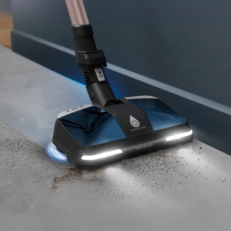 Czarna i niebieska głowica odkurzacza ze światłami czyszcząca brudną podłogę. Uchwyt jest różowy i czarny. Ściana jest niebieska.