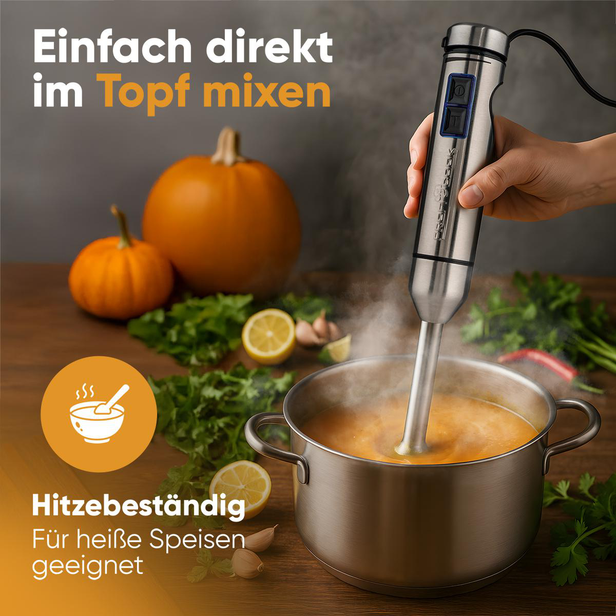 Eine Hand hält einen Mixer, der Suppe in einem Topf verrührt. Kürbisse, Zitronen und Kräuter liegen auf dem Holztisch.