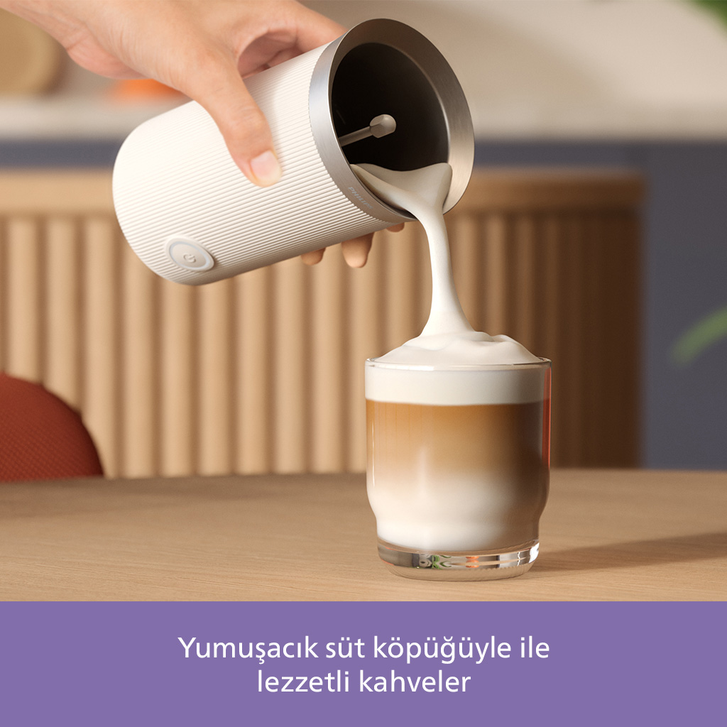 Bir el, bir bardak kahveye köpüklü süt döküyor. Süt köpürtücü beyaz, kahve katmanlı. Sahne ahşap bir masa üzerinde.