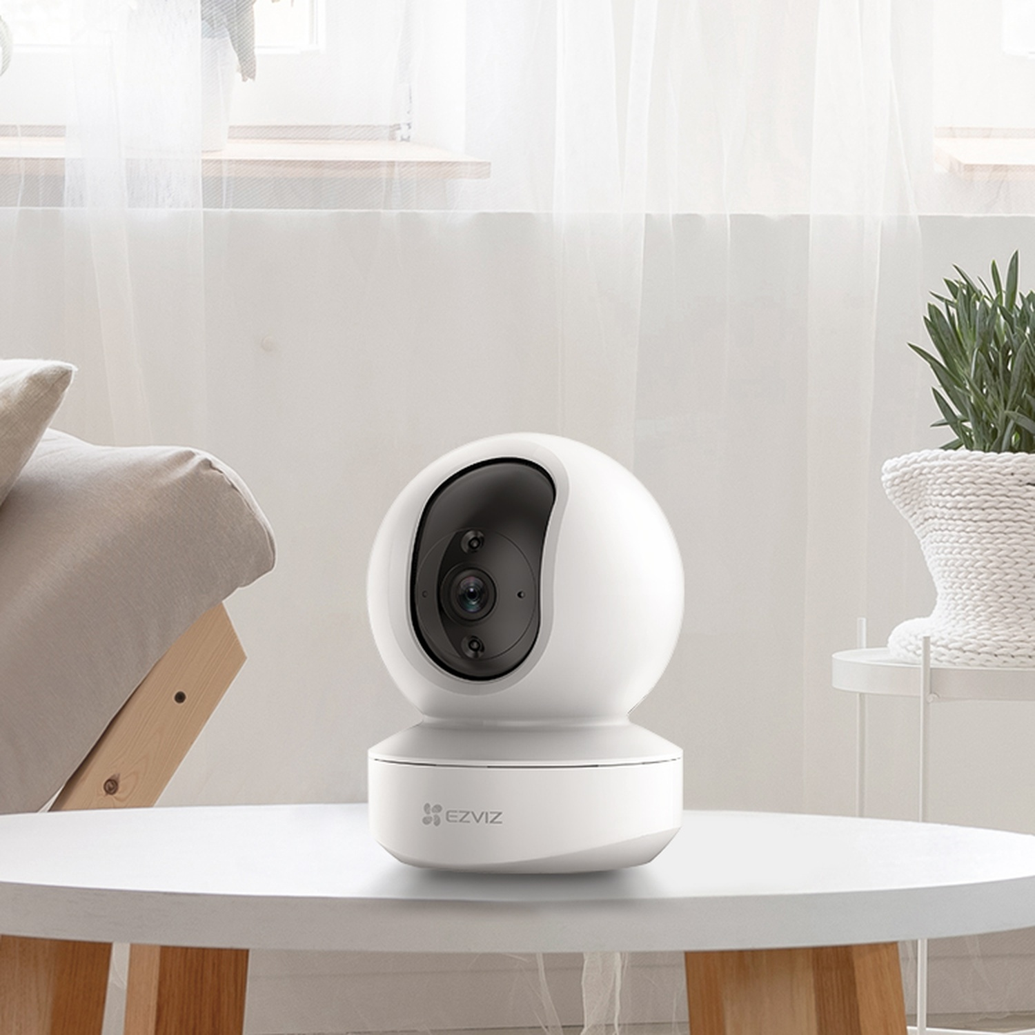 Una telecamera di sicurezza EZVIZ bianca è su un tavolo bianco. Un letto e una pianta sono sullo sfondo, vicino a una finestra.