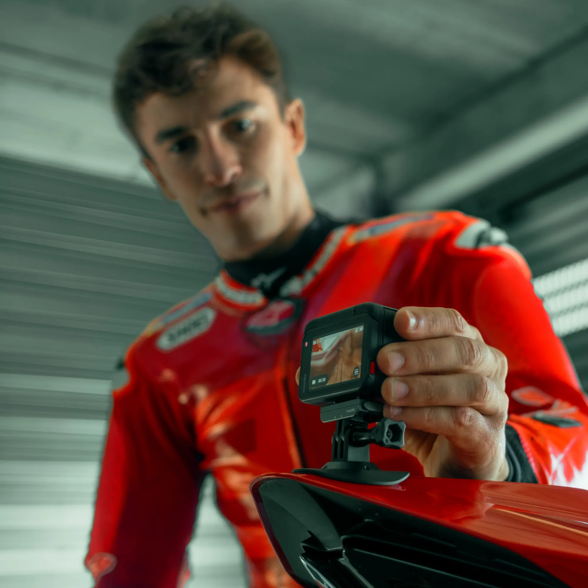 Ein Mann in rotem Rennanzug hält eine Kamera. Die Kamera ist auf einem roten Motorrad montiert. Der Hintergrund ist eine Garage.