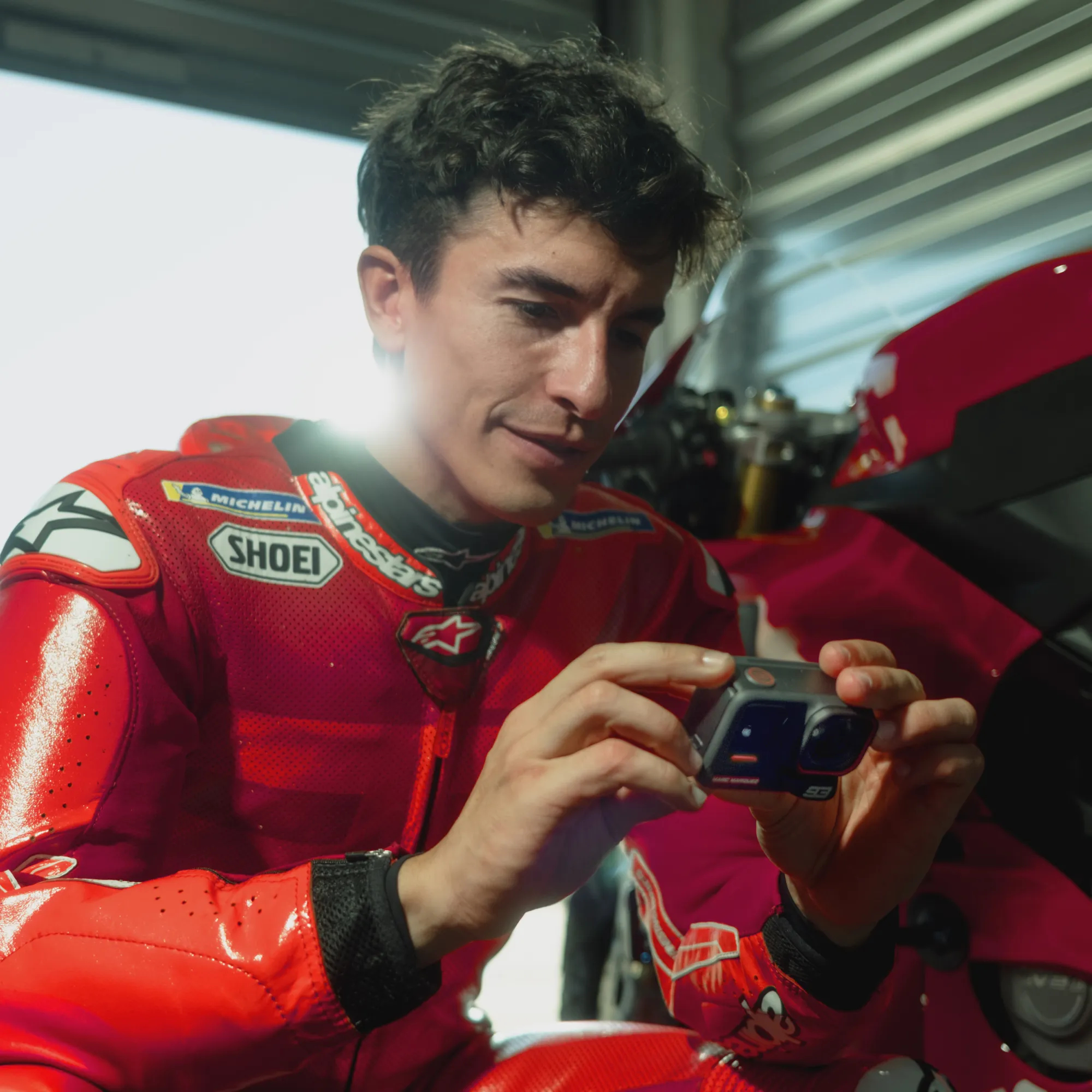 Ein Mann in rotem Rennanzug untersucht eine Kamera. Im Hintergrund ist ein Motorrad zu sehen. Die Szene findet drinnen statt.