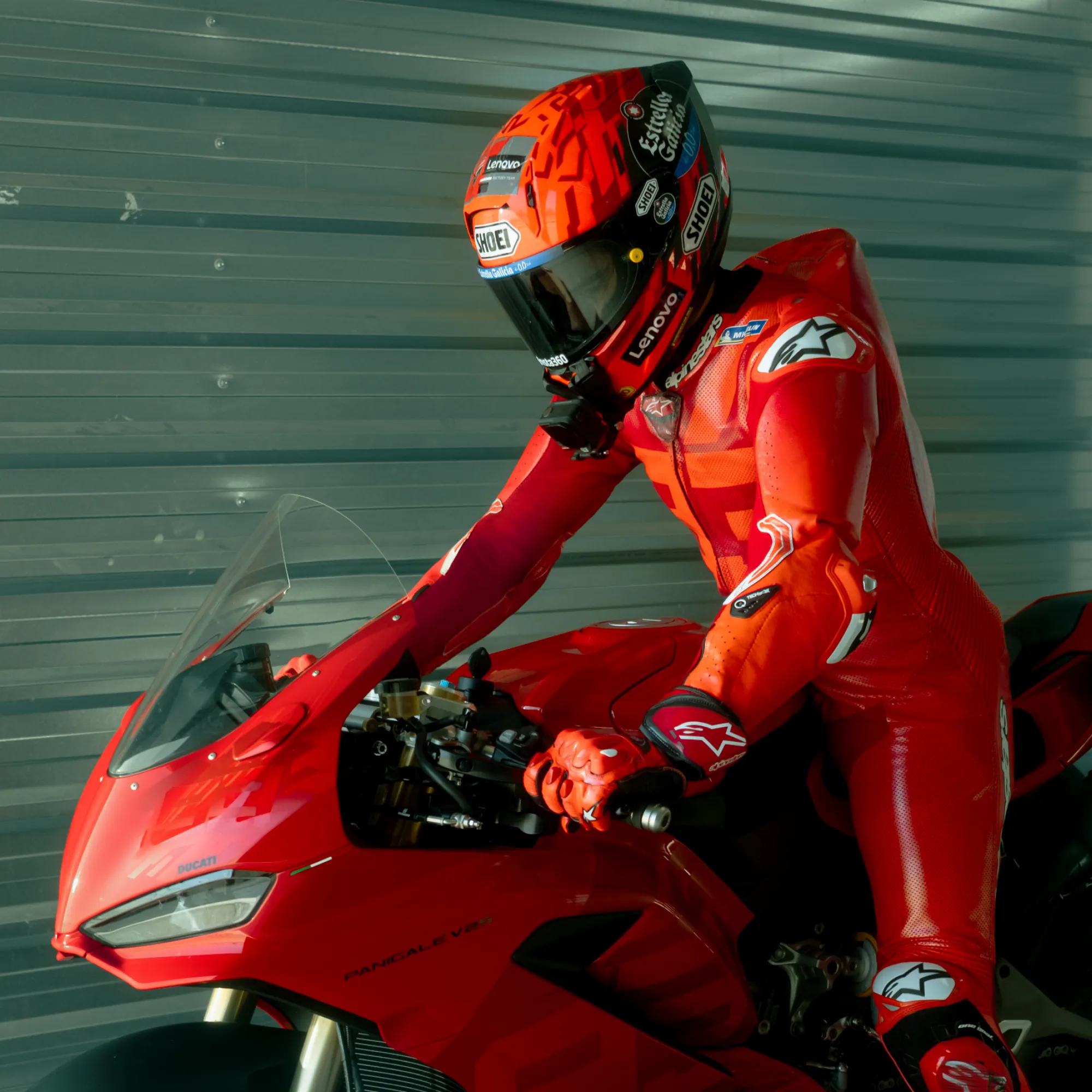 Eine Person in rotem Rennanzug und Helm sitzt auf einem roten Ducati Panigale Motorrad. Der Hintergrund ist eine graue Wand.