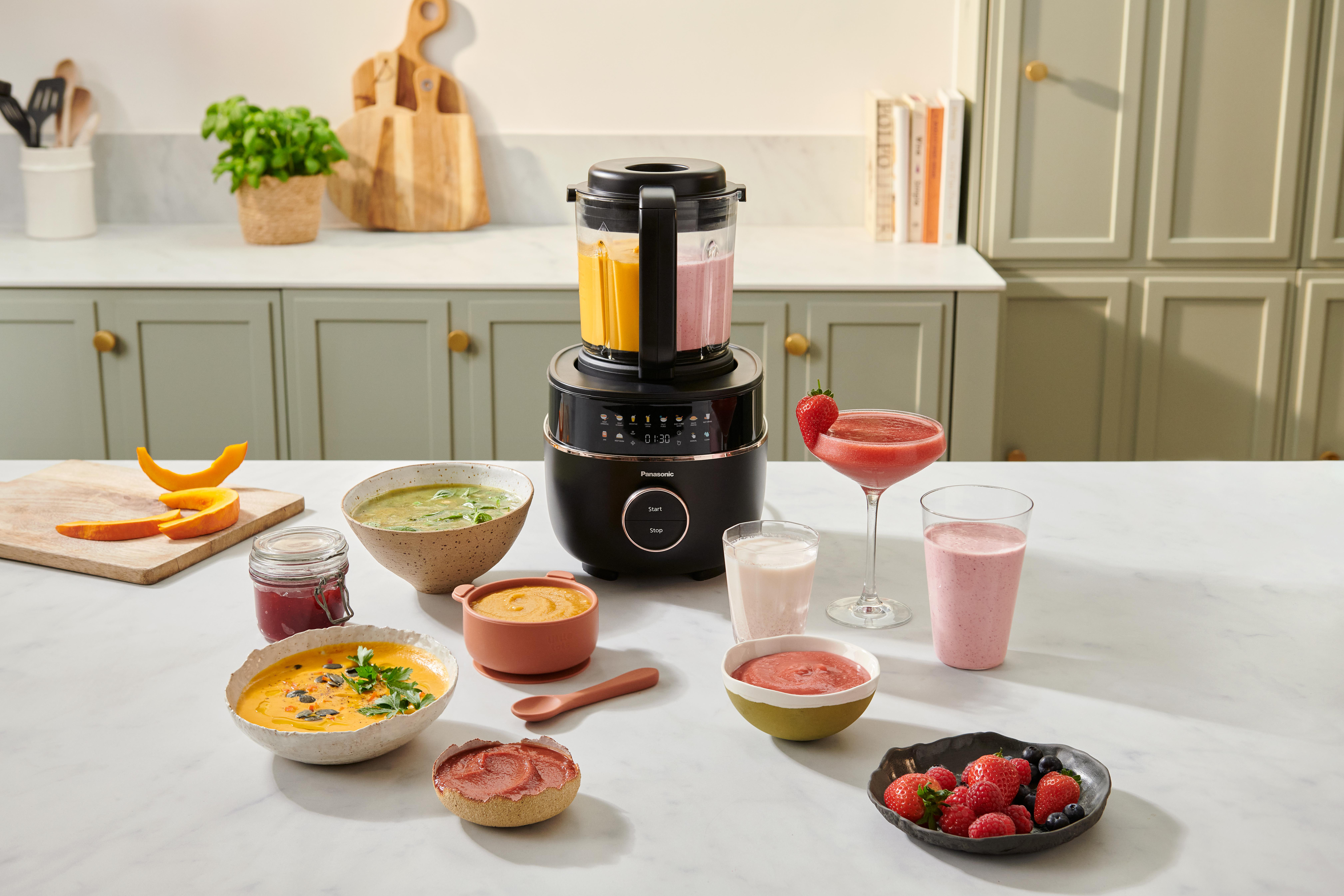Ein Mixer mit gelben und rosa Flüssigkeiten, verschiedene Suppen, Smoothies, Früchte und ein Schneidebrett mit Kürbisscheiben auf einer Theke.
