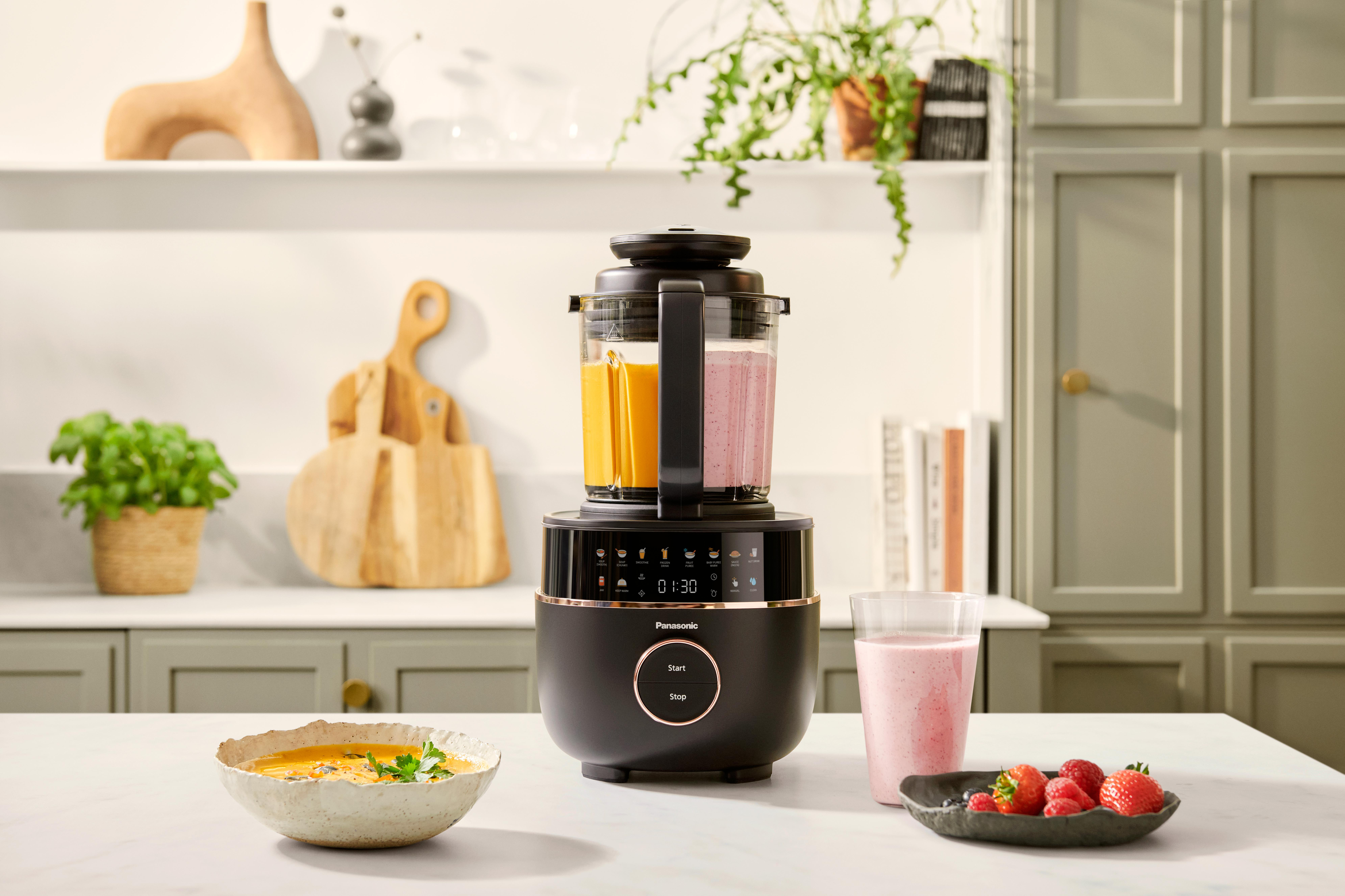 Ein Mixer mit orangefarbenen und rosa Flüssigkeiten. Suppe und Beeren sind vorne. Pflanzen und Schränke sind im Hintergrund.