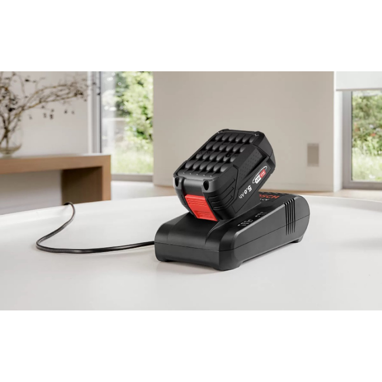 Batterie de perceuse sans fil noire et rouge sur un chargeur noir. Le chargeur est sur une table blanche. L'arrière-plan montre une fenêtre.