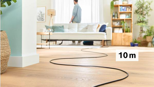 Wohnzimmer. Ein schwarzes Kabel schlängelt sich über den Holzboden. Ein Mann steht an einem weißen Sofa.