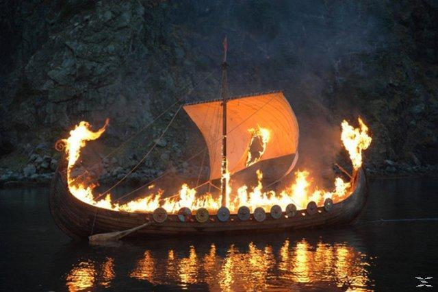 Ein brennendes Wikingerschiff auf dem Wasser. Flammen verschlingen das Holzschiff und spiegeln sich im dunklen Wasser. Im Hintergrund ein felsiger Abhang.