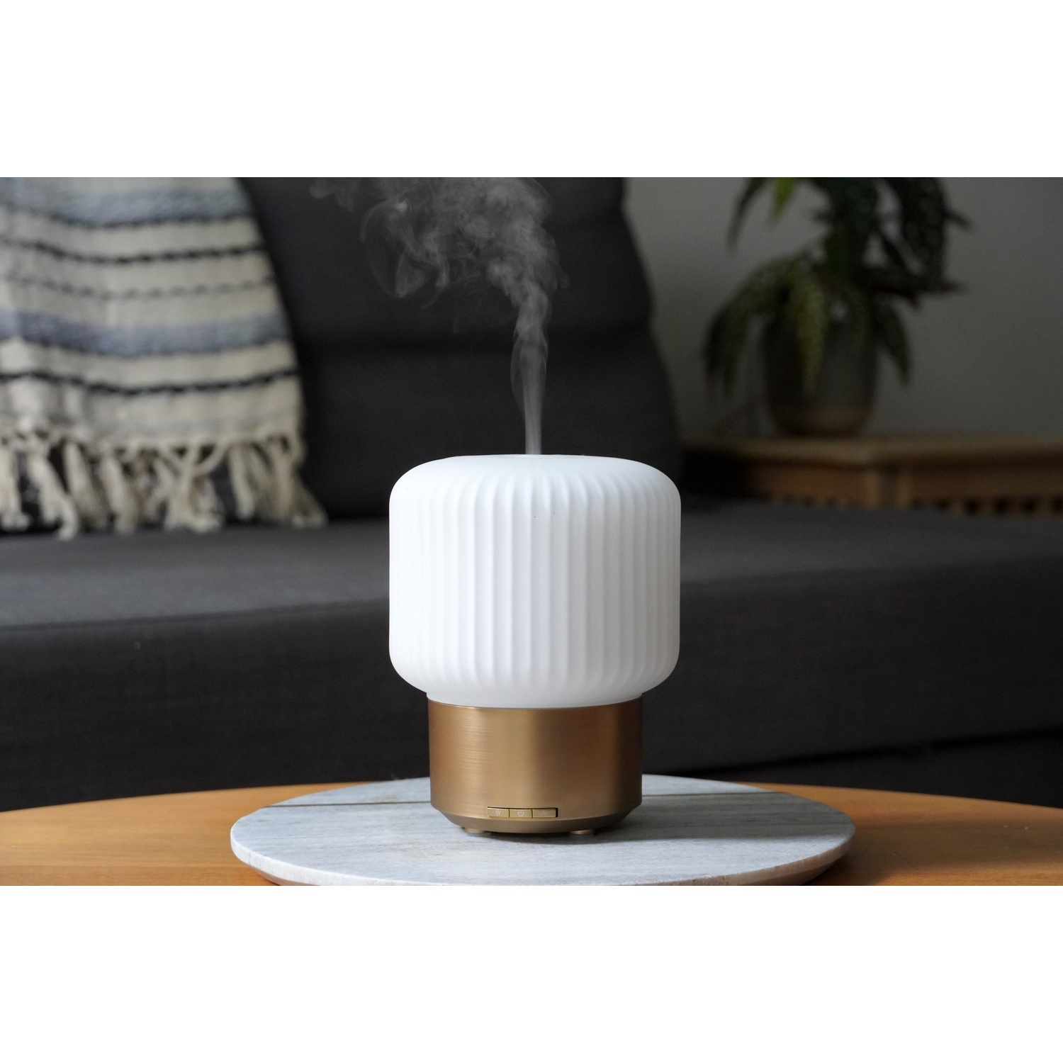 A white and gold aroma diffuser on a marble coaster. Smoke rises from the diffuser. A sofa and plants are in the background.