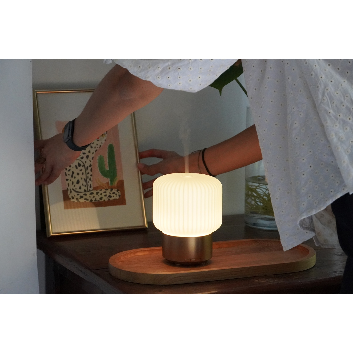 A person placing a picture frame on a wooden table. There's a glowing lamp and a wooden tray. The person wears a white shirt.