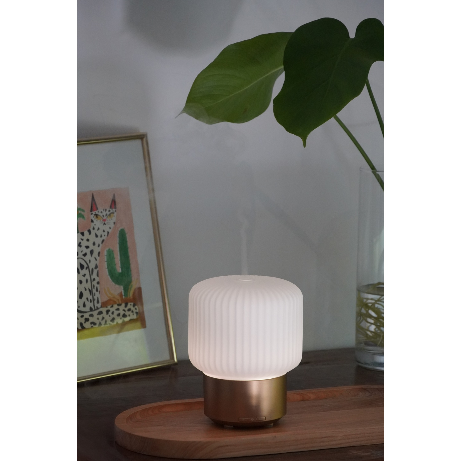 A white and gold diffuser sits on a wooden tray. A framed picture and green plant are in the background.