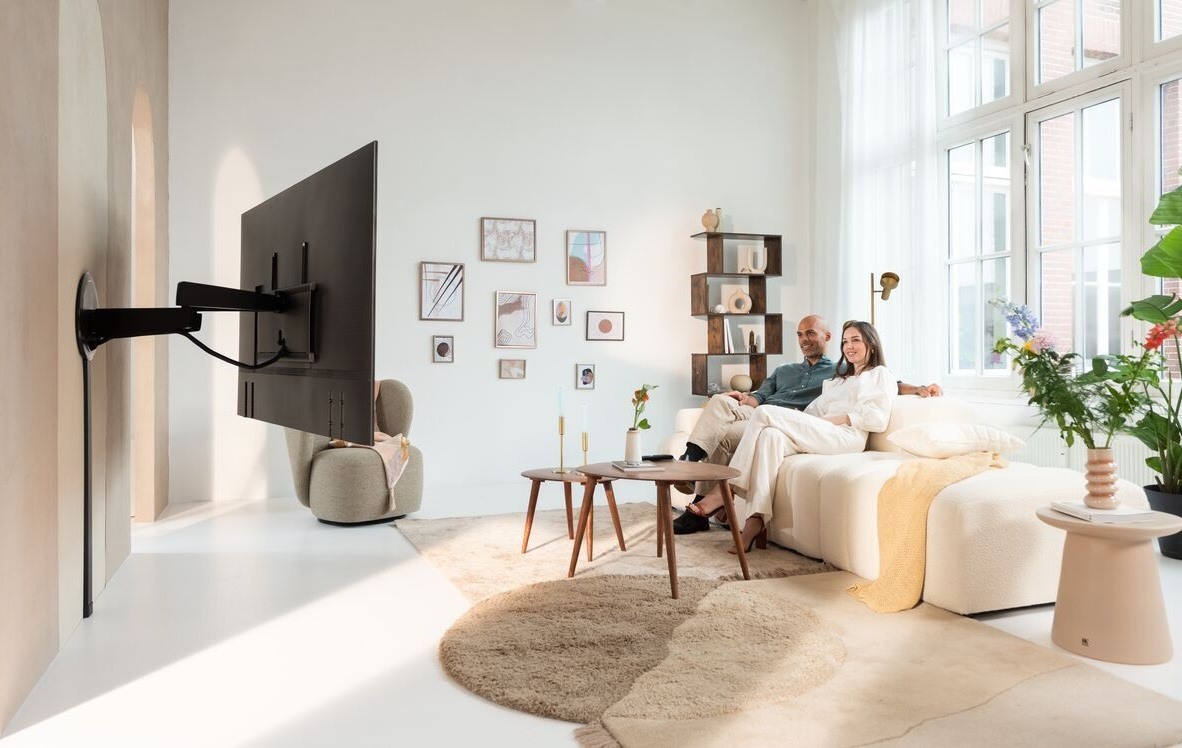 Para siedzi na kanapie. Telewizor jest zamontowany na ścianie. Dywan i dwa stoliki kawowe znajdują się na podłodze.