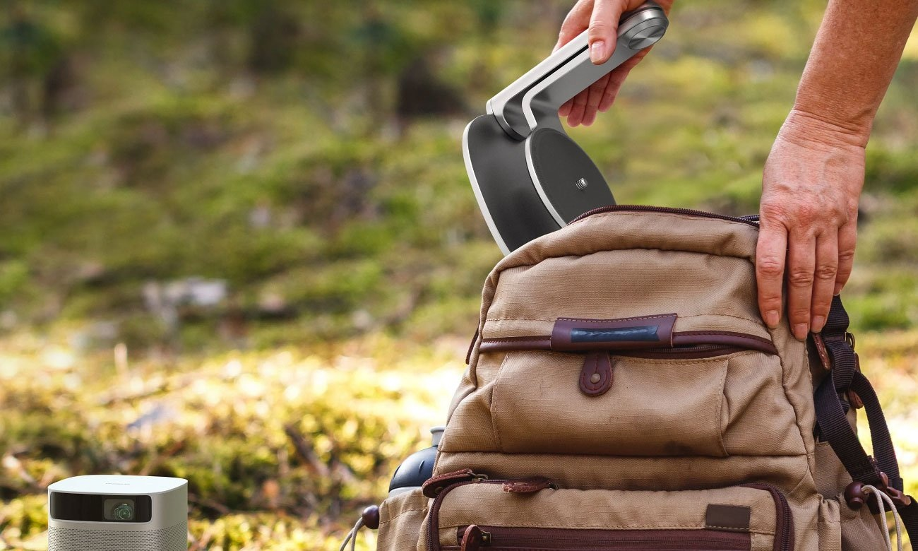 Osoba umieszcza czarno-srebrne urządzenie w brązowym plecaku. W pobliżu, w zielonym otoczeniu, znajduje się projektor.