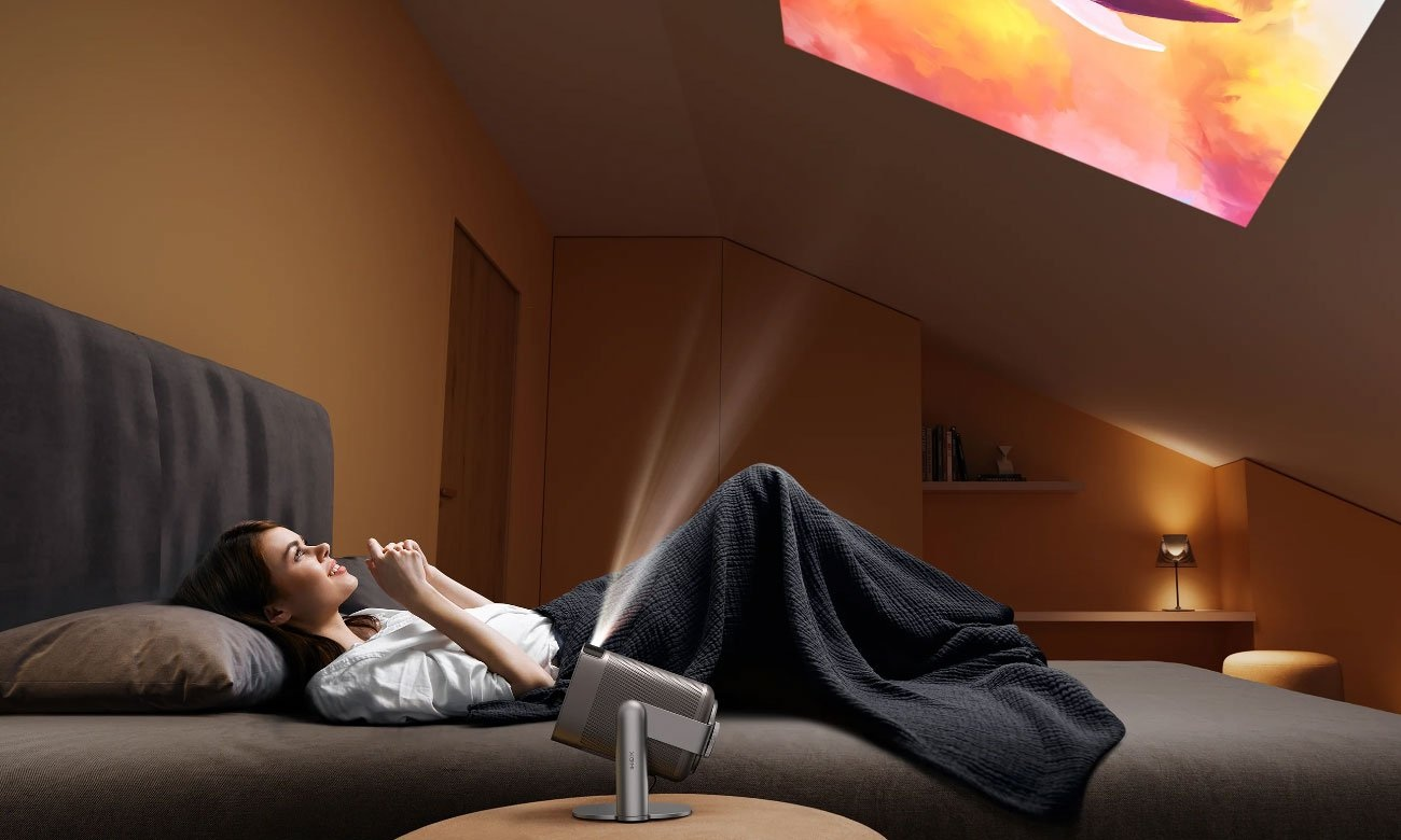 Kobieta leży na łóżku, oglądając wyświetlany obraz. W pobliżu stoi projektor. Pokój jest słabo oświetlony.