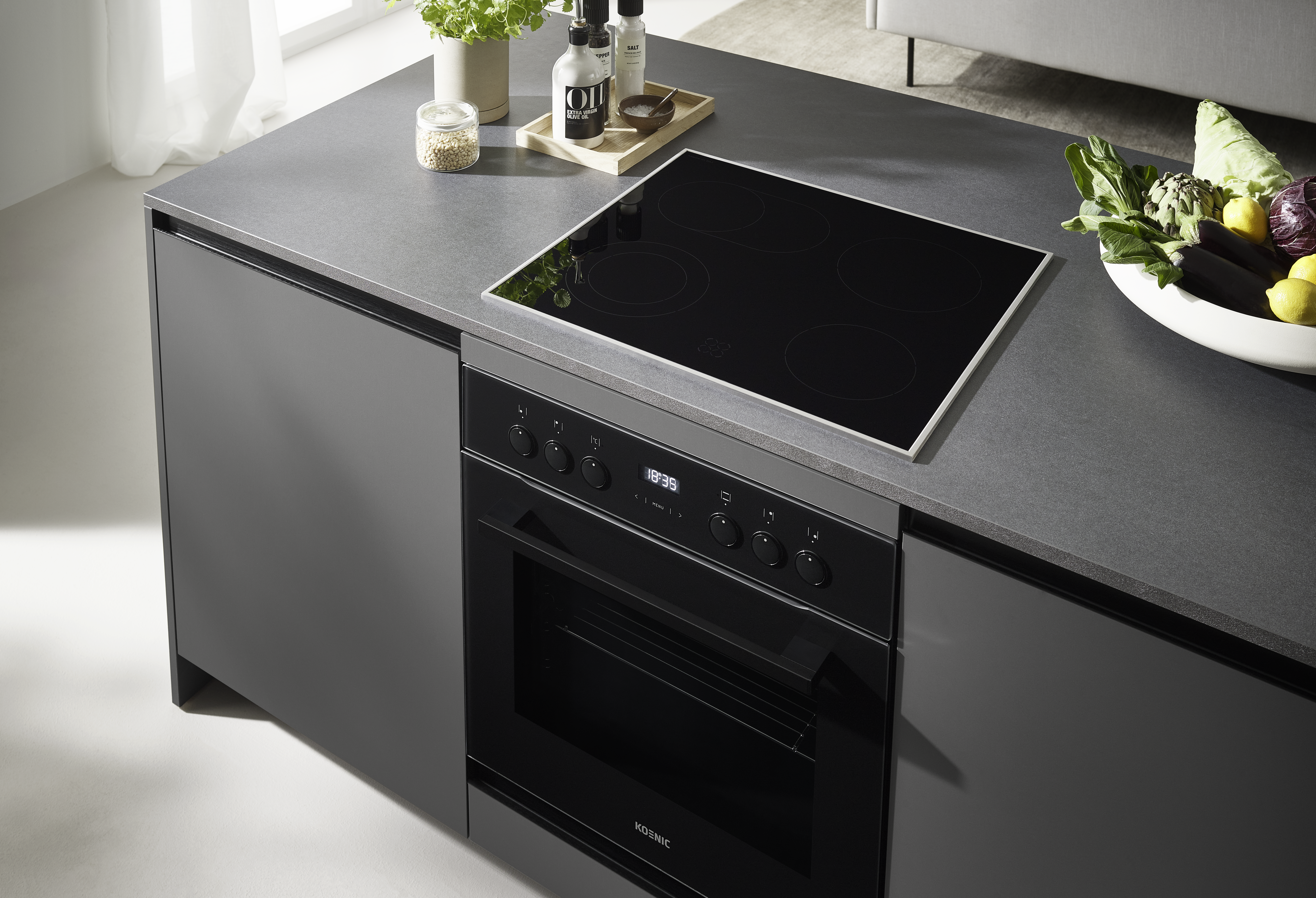 Moderne Küche mit schwarzem Kochfeld, Backofen und Arbeitsplatte. Gemüse und Zitronen in einer weißen Schüssel. Andere Zutaten auf der Theke.