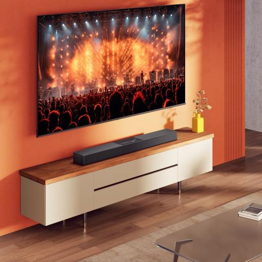 Egy nagy TV koncertjelenetet jelenít meg egy soundbar felett. Egy szekrény tartja a soundbart és egy sárga vázát. Narancssárga fal.