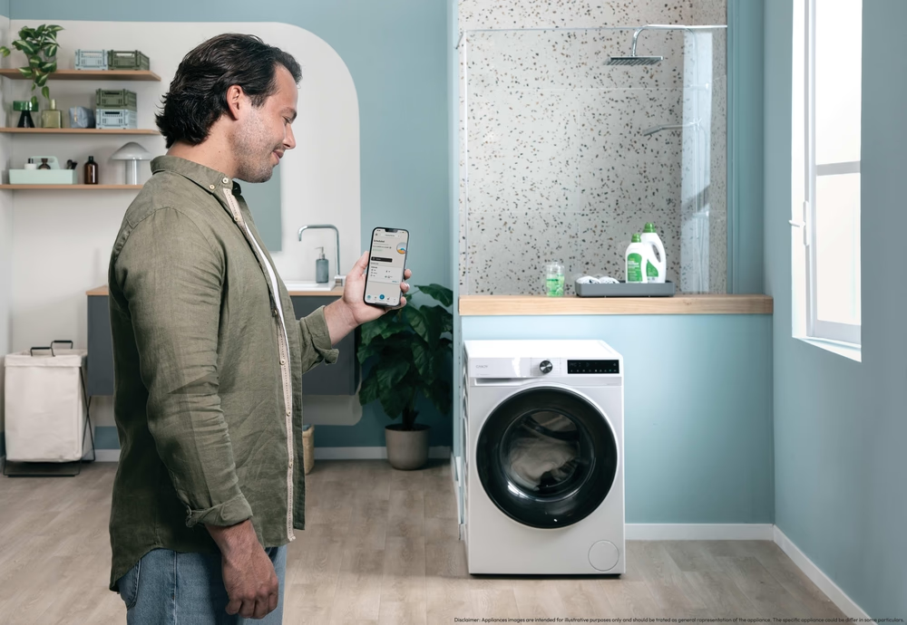 Egy férfi a telefonját nézi egy fürdőszobában. Előtte egy mosógép áll. A háttérben zuhanyzó, mosdókagyló és polcok láthatók.
