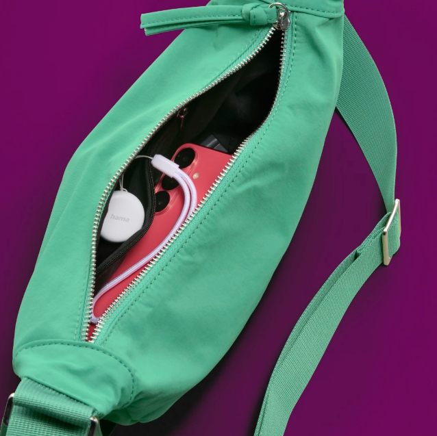 Offene grüne Tasche mit Telefon, weißem runden Objekt und Ohrhörern. Lila Hintergrund. Reißverschluss ist offen.