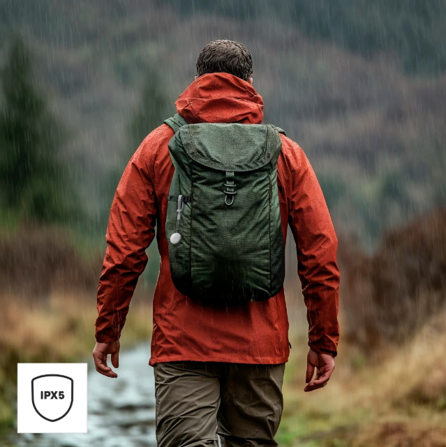 Eine Person mit roter Jacke und grünem Rucksack geht im Regen. Der Hintergrund zeigt einen verschwommenen Wald. IPX5-Symbol sichtbar.
