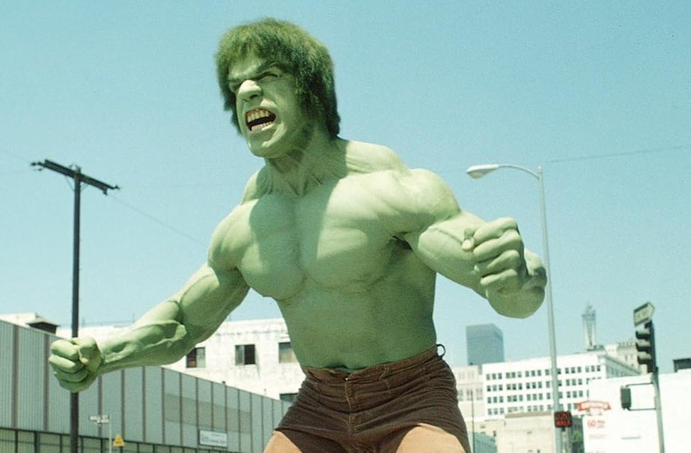 Der Hulk in Aktion in einer Stadt. Er ist grün, muskulös und trägt braune Shorts. Gebäude und blauer Himmel sind sichtbar.